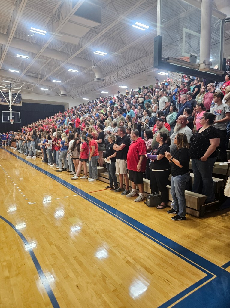 A large crowd of people standing in a gymnasium, with many in red and blue clothing, showing community support during a school assembly.