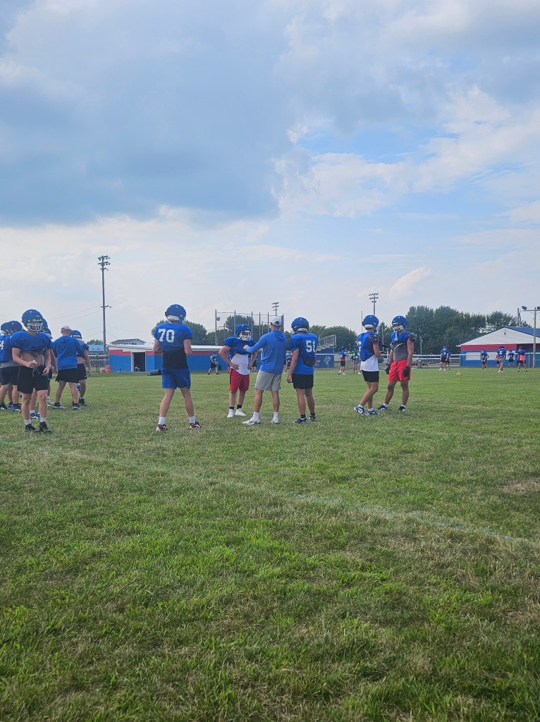 A football practice session with players in blue uniforms gathered on a grassy field, discussing strategies with a coach in a white shirt.