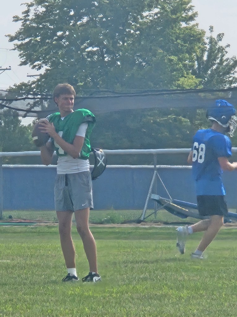 A high school football player in a green practice jersey prepares to throw a football during practice, with another player in a blue jersey walking in the background.