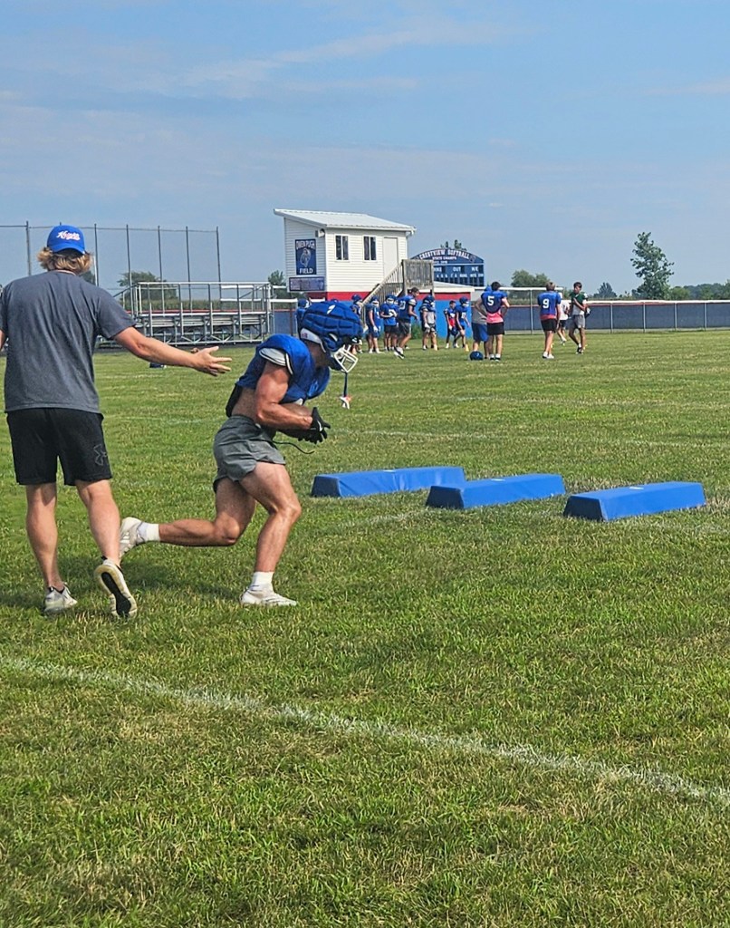 A football player practices running drills on a grassy field, guided by a coach. Blue training equipment is visible in the background along with other players in practice.