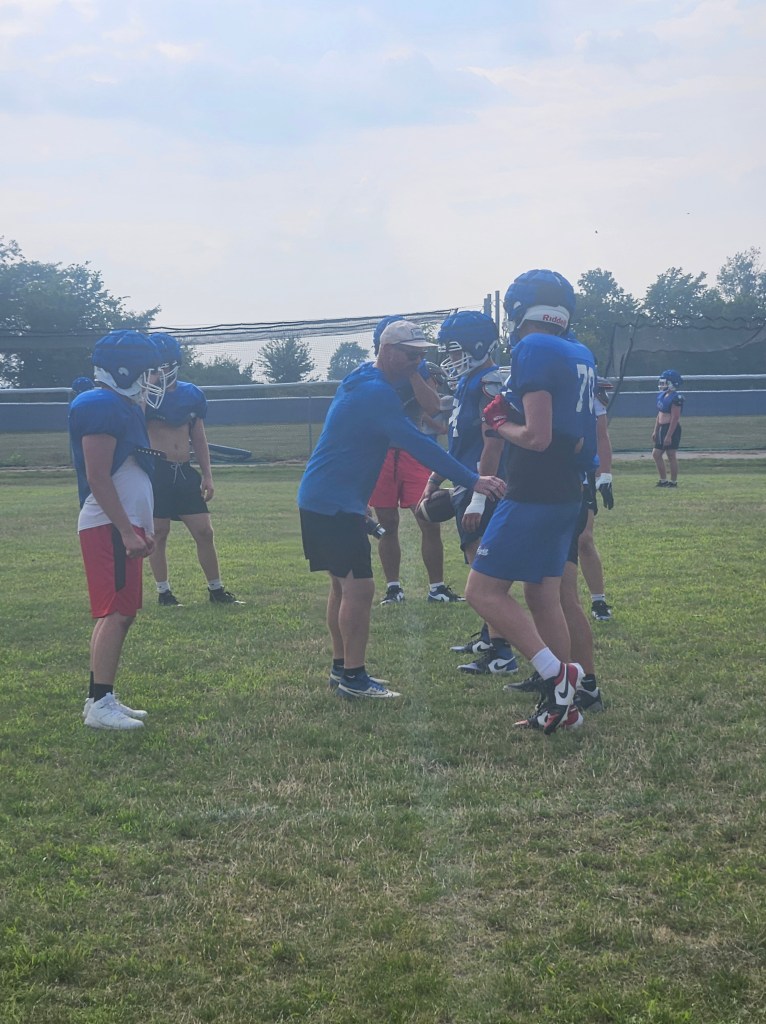 A football coach instructs players during a practice session on a grassy field. Several athletes in football gear gather around, attentively listening and engaged in discussion.