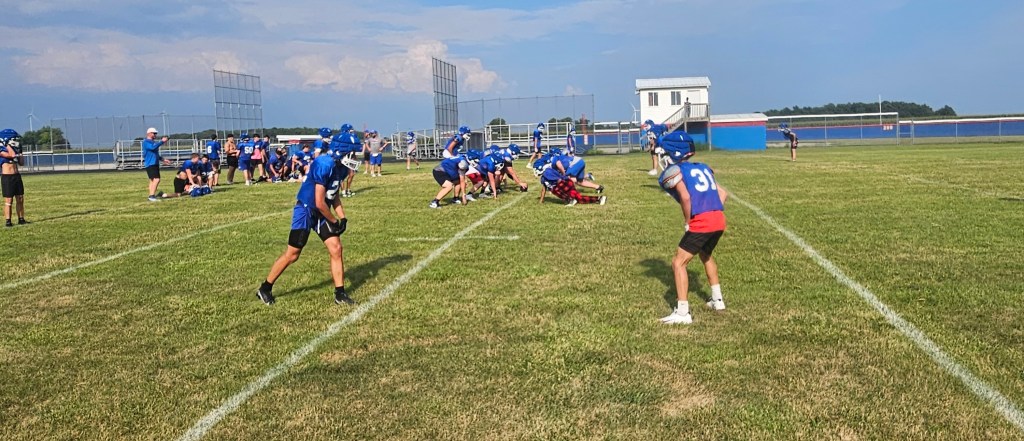 High school football practice scene with players in blue jerseys on a grassy field, some in position preparing for drills.