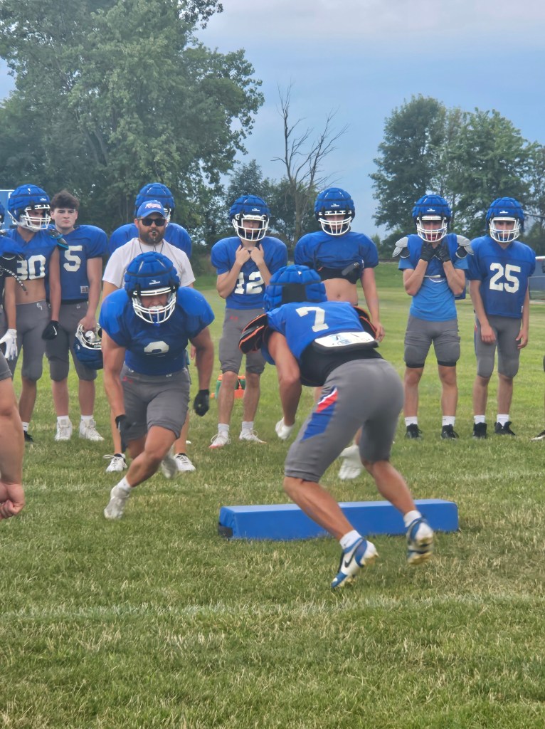 High school football practice featuring players in blue and gray uniforms, with a coach observing as they simulate drills on a grassy field.