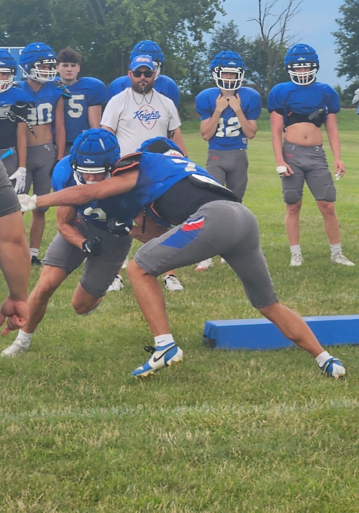 Two high school football players are practicing a tackling drill on a grassy field, with coaches and teammates observing in the background.