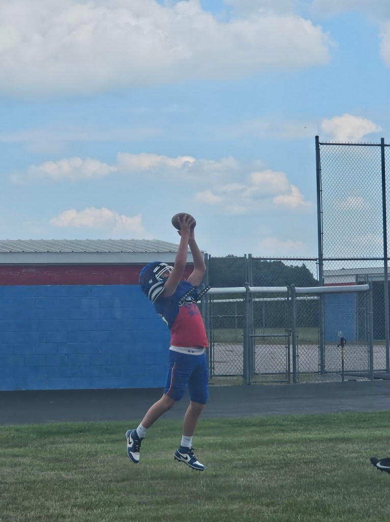 A young football player jumps to catch a pass during practice, wearing a helmet and blue and red uniform against a clear sky with clouds.