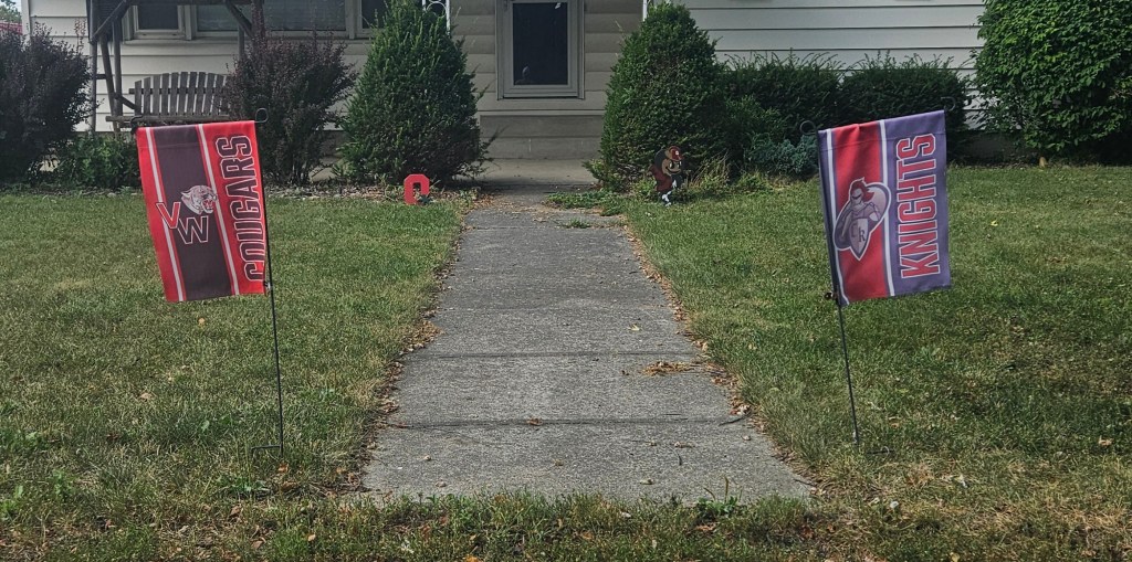 Flags representing the Van Wert Cougars and Crestview Knights football teams are displayed on a grassy lawn, signaling community support for the local high school rivalry.
