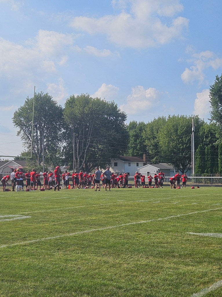 Football players in red uniforms practicing on a green field under a blue sky with scattered clouds.