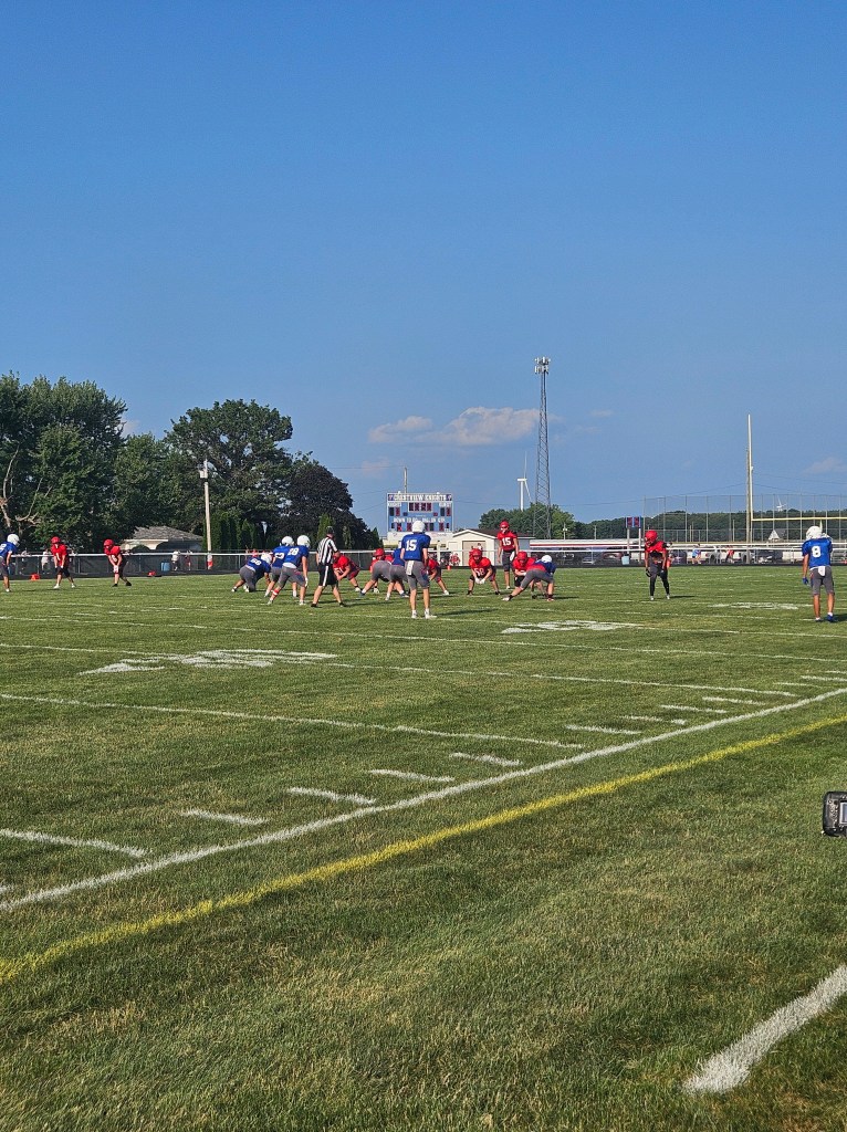 A high school football scrimmage in Van Wert County, Ohio, with players from two teams lined up on the field. The scene shows a clear blue sky, green grass, and the scoreboard in the background.