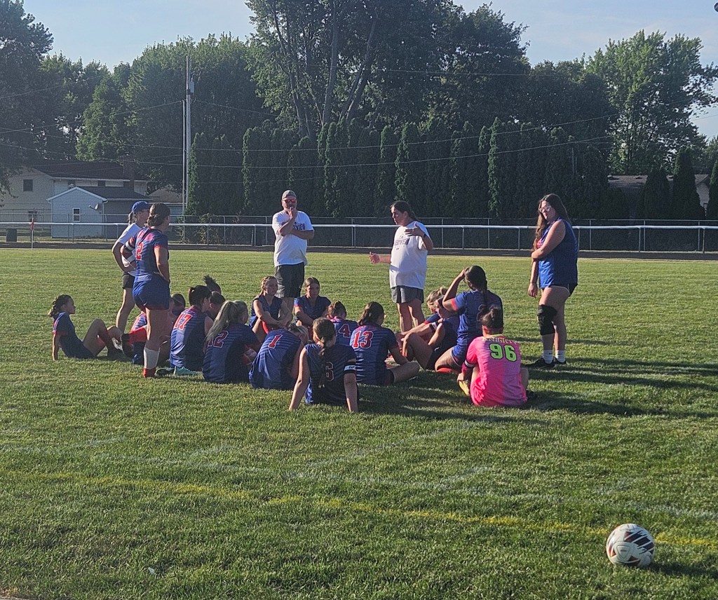 A group of Crestview Lady Knights girls soccer team members gathered for a discussion on the field, with their coach giving instructions while some players sit and others stand around.