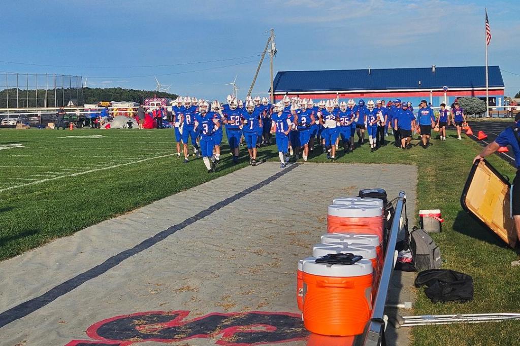 Crestview Knights football team walking onto the field before a game, wearing blue uniforms, with coaches and staff in the background.