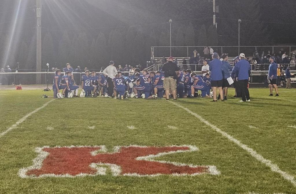 A group of Crestview Knights football players and coaches gathered on the field, discussing strategies during a night game.