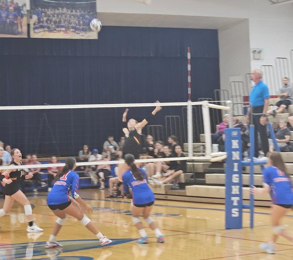 A volleyball match in progress with players from both teams in action. The Crestview Lady Knights are in black uniforms, while the New Bremen Cardinals are in blue uniforms. A player from Crestview is jumping to hit the ball over the net.