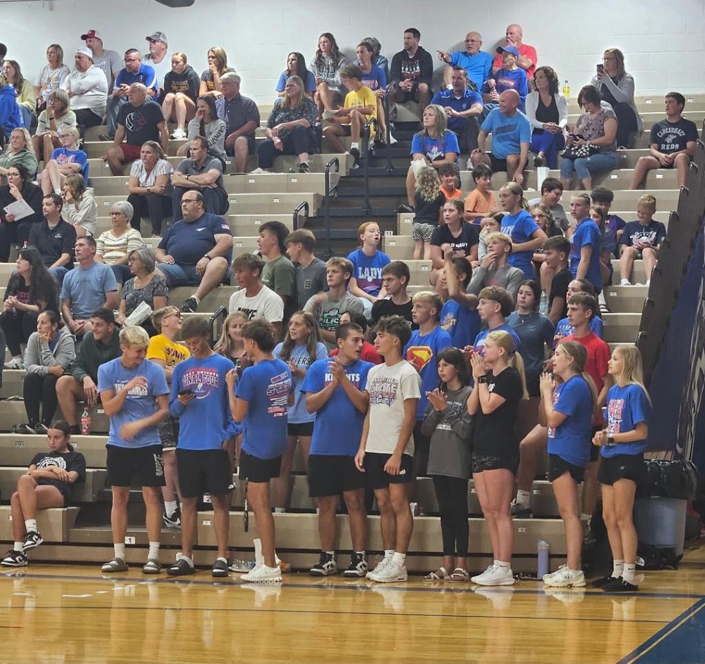 A large group of students and fans cheering in the stands during a high school volleyball game, showcasing team spirit with school colors and various expressions of excitement.