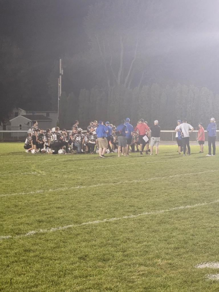 Football team huddled together on the field during a night game, surrounded by coaches and support staff.