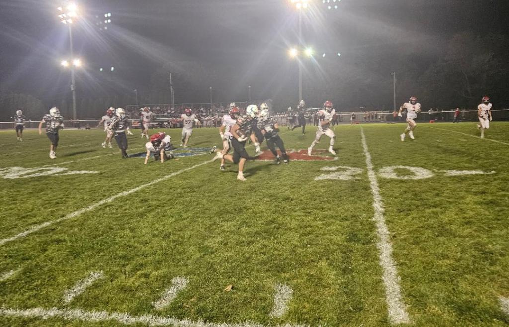 A football play in progress during a night game, with players in dark and light uniforms on the field, bright stadium lights illuminating the scene.