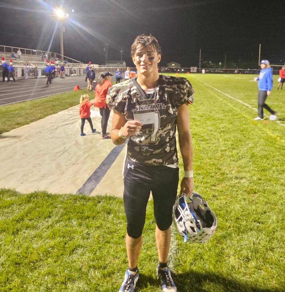 A football player from the Crestview Knights stands on the sidelines after a game, wearing a camouflage jersey and holding a ticket while smiling for the camera.