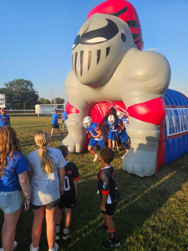 A group of children excitedly watches as football players exit an inflatable knight tunnel at a field during warm late summer evening.