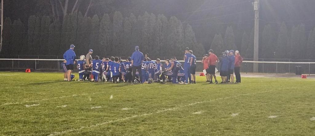 A group of football players in blue jerseys huddled together on the field during a nighttime practice, surrounded by coaches and staff.