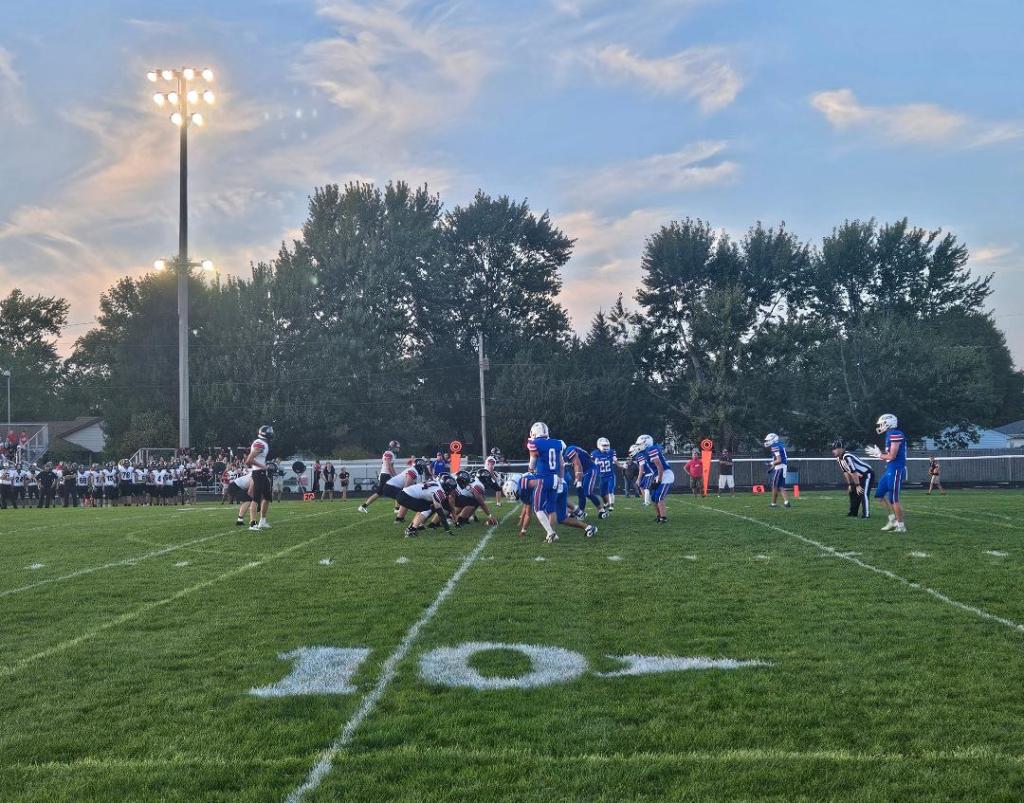 A youth football game in Convoy, Ohio, featuring players in blue and black uniforms on the field during a warm summer evening, with spectators in the background.