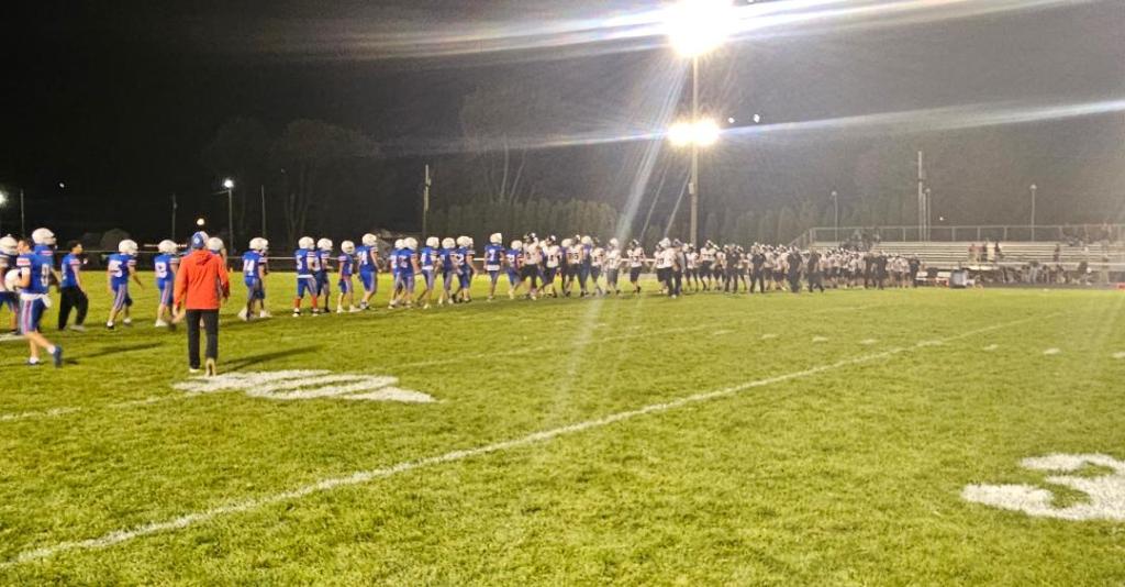 Two football teams shaking hands at the end of a night game on a well-lit field.
