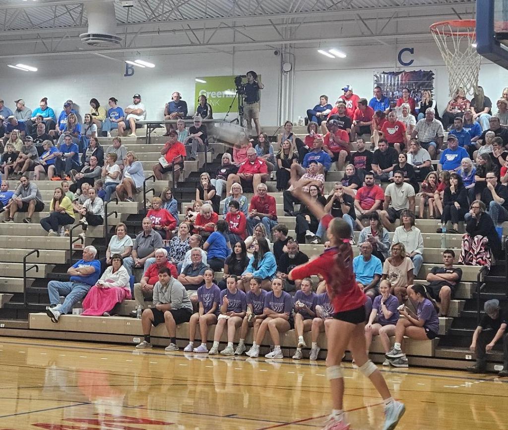 A packed gymnasium filled with fans watching a volleyball match, with a player preparing to serve in the foreground.