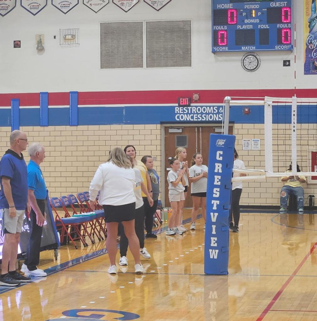 Players and coaches gathered in a gymnasium before a volleyball match between Crestview and Lincolnview, with the scoreboard showing 0-0.