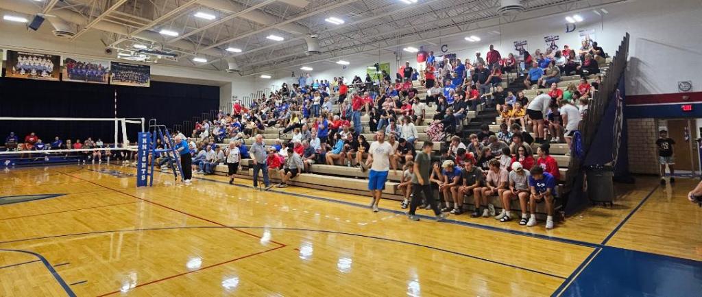 A crowded gymnasium filled with spectators watching a volleyball match, featuring bleachers on both sides and a volleyball net in the foreground.