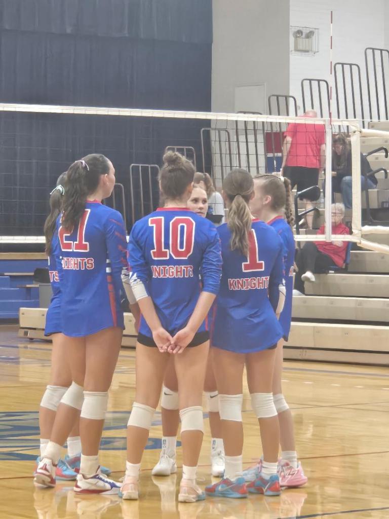 Crestview Lady Knights volleyball team in a huddle during a match, wearing blue and red uniforms.