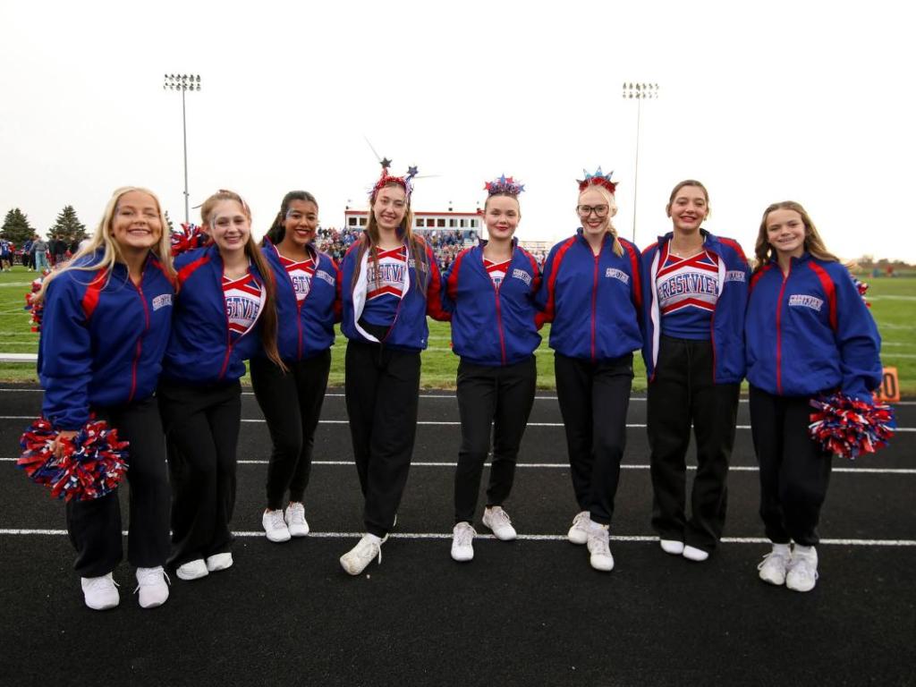 A group of cheerleaders in Crestview Knights athletic attire, posing on the sidelines of a football field, with a crowd in the background.