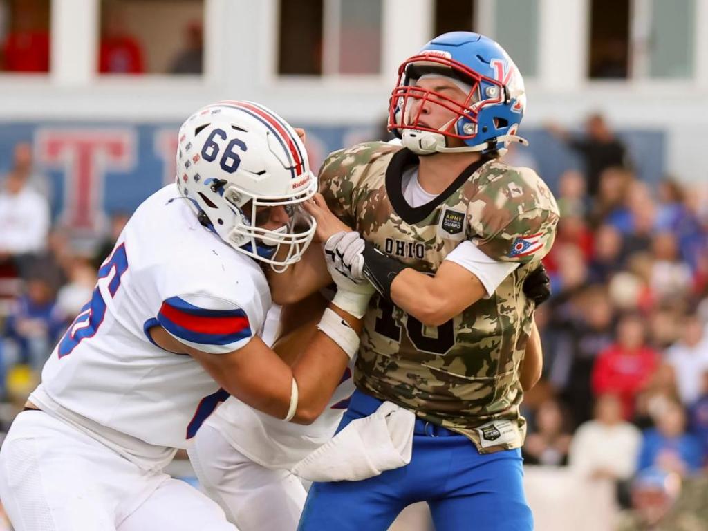 A high school football player in camo jersey is tackled by an opponent during a game.