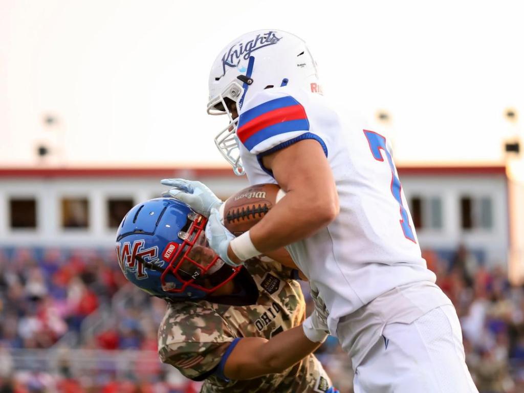 A football player from the Crestview Knights team tackles an opposing player from the Wayne Trace Raiders during a high school football game under a setting sun.