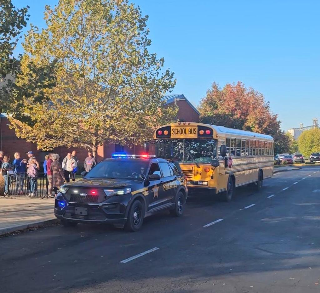 A police escort vehicle with flashing lights is positioned in front of a yellow school bus, while a crowd of people gathers on the sidewalk, set against a backdrop of trees with autumn foliage.