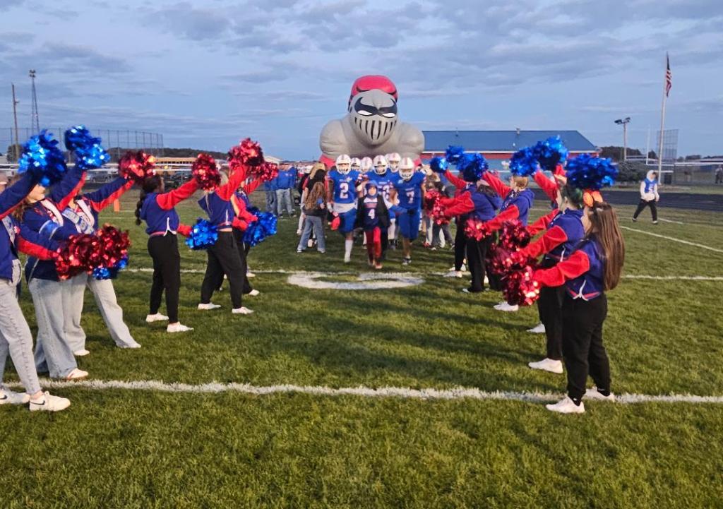 Cheerleaders with pom-poms welcoming the football team onto the field against a backdrop of a gray sky and stadium.