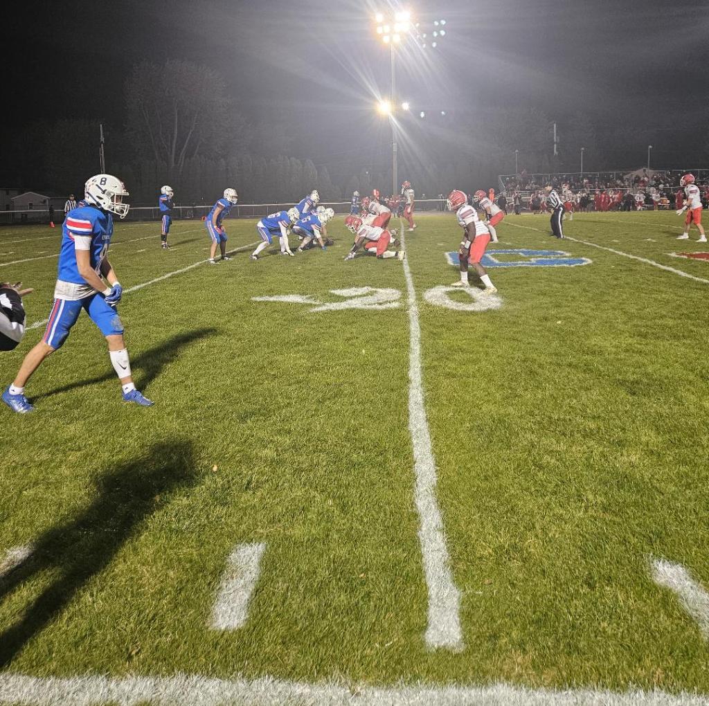 A high school football game in progress, with players on the field wearing blue and red uniforms, against a backdrop of stadium lights.