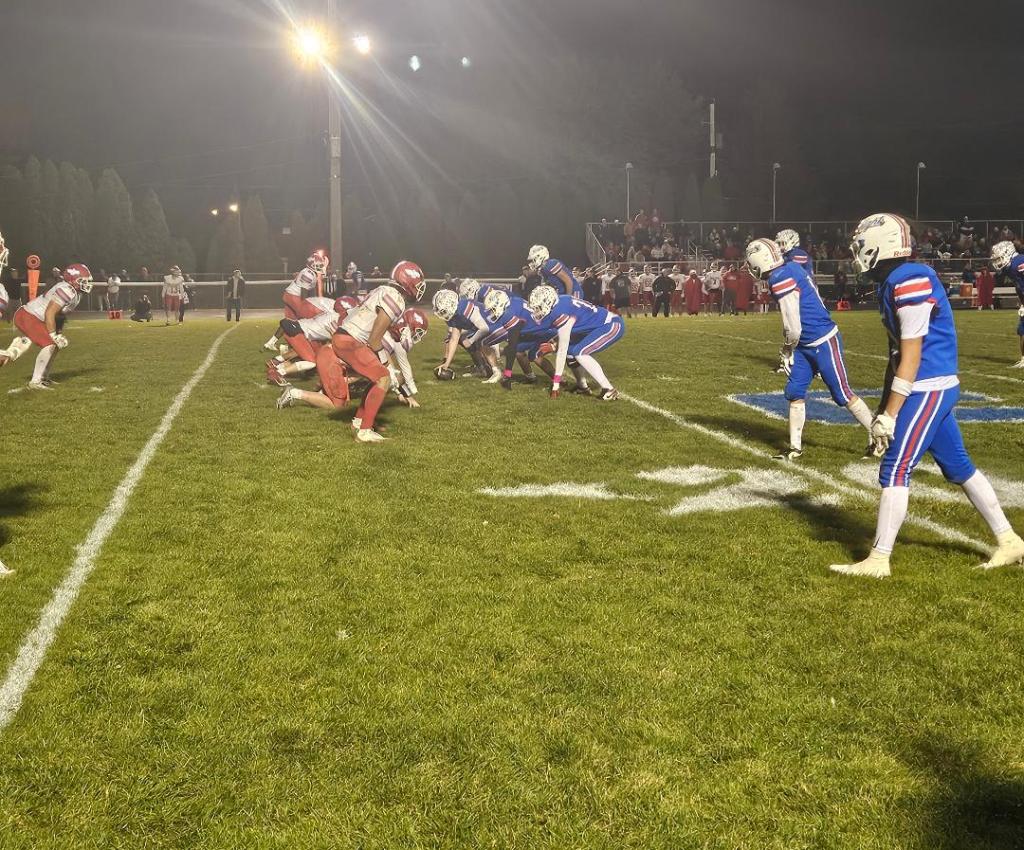 Two football teams lined up on a grass field, with players in red and blue uniforms ready for a play during a night game.