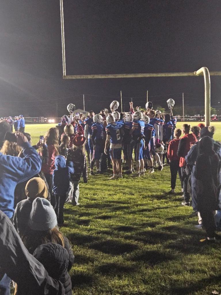 A high school football team gathers together under the goalpost, celebrating and honoring their seniors after a night game, with spectators watching from the sidelines.