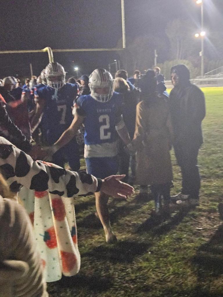 A group of football players in blue jerseys walking hand-in-hand with a crowd of supporters after a game under night lights.