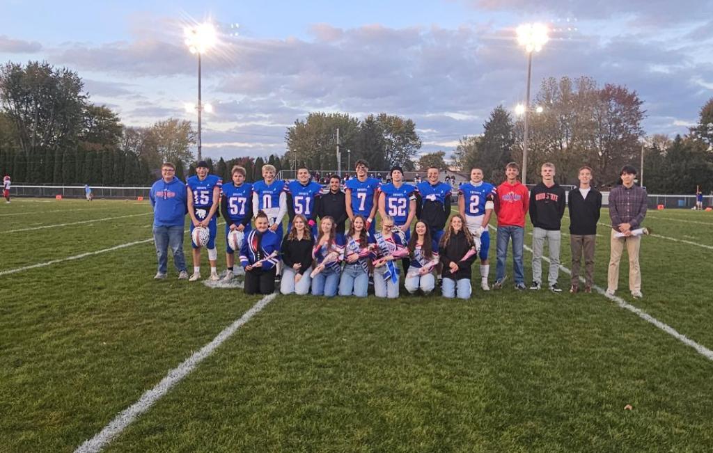 A group of high school seniors posing together on a football field during Senior Night, wearing team jerseys and holding flowers, with lights shining in the background.