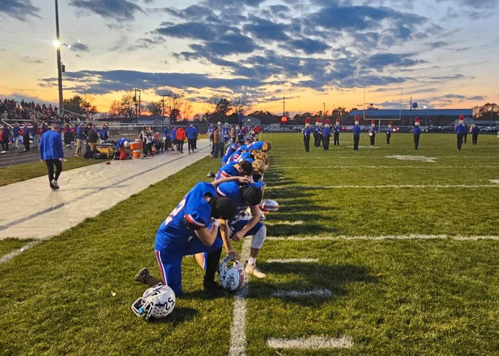 A group of football players kneeling on the sidelines during a sunset, preparing for a game. The sky is filled with orange and purple hues, and a crowd can be seen in the background.