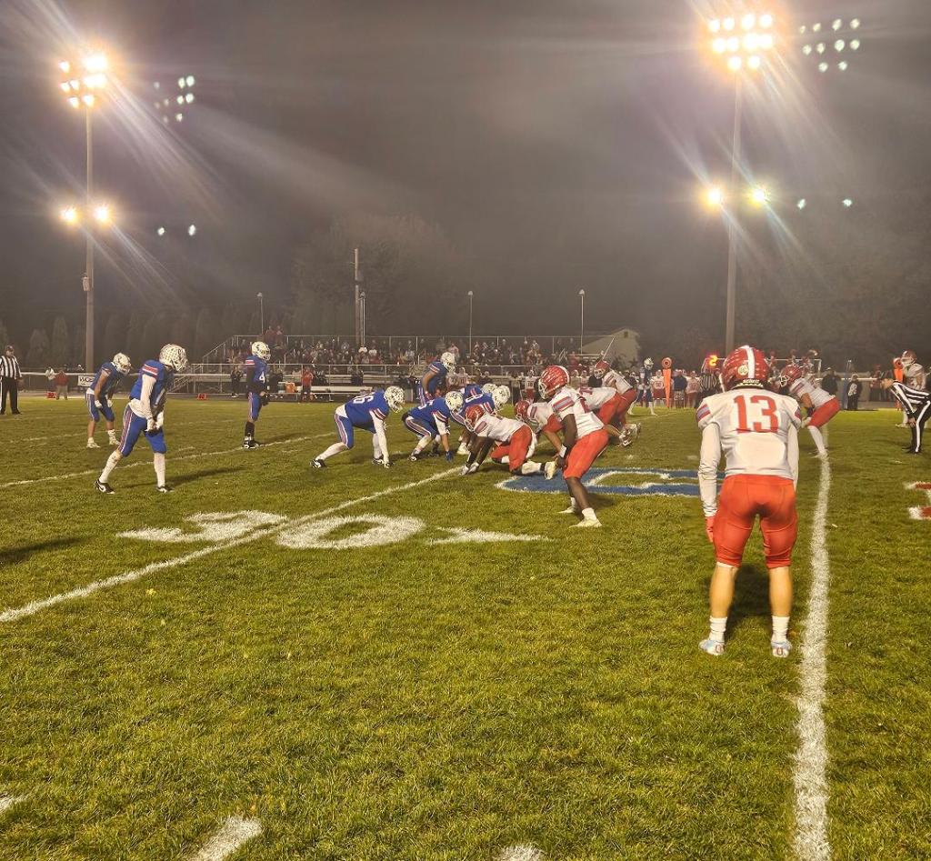A high school football game in progress under bright stadium lights, with players from two teams lined up for a play on the field.