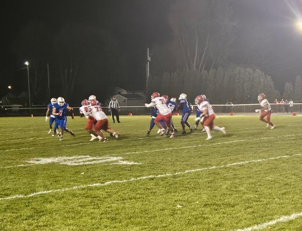 A high school football game in progress, featuring players in red and blue uniforms on a grassy field under night lights.