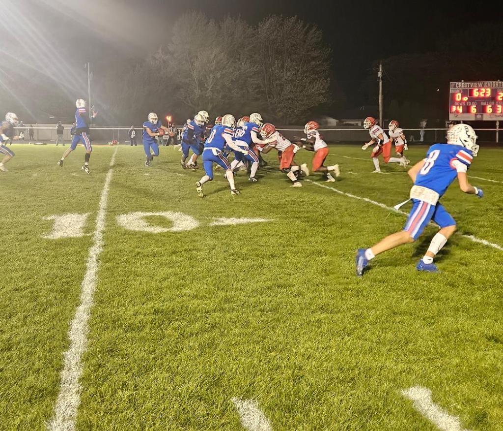 A high school football game in progress, featuring players in blue and white jerseys against red and white jerseys, on a well-maintained grass field under bright stadium lights.