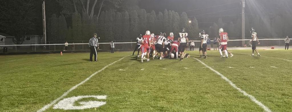 A competitive moment during a high school football game, with players from both teams engaged in a pile-up on the field under night lights.