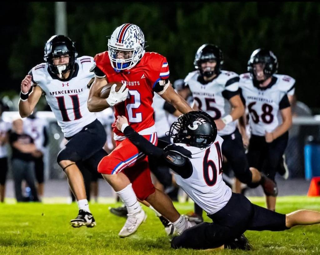A football player wearing a red jersey with the number 2 is evading a tackle from a player in a black jersey during a nighttime game, while additional players from both teams are visible in the background.