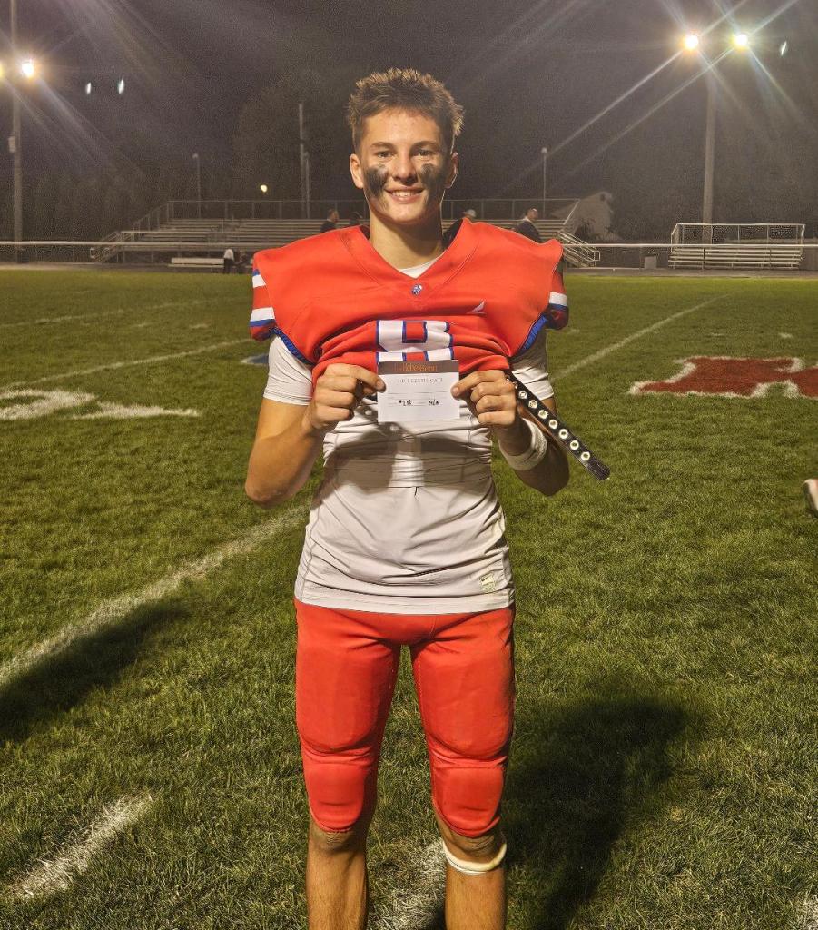 A football player wearing a red jersey and pants stands on the field, smiling and holding up an award certificate.