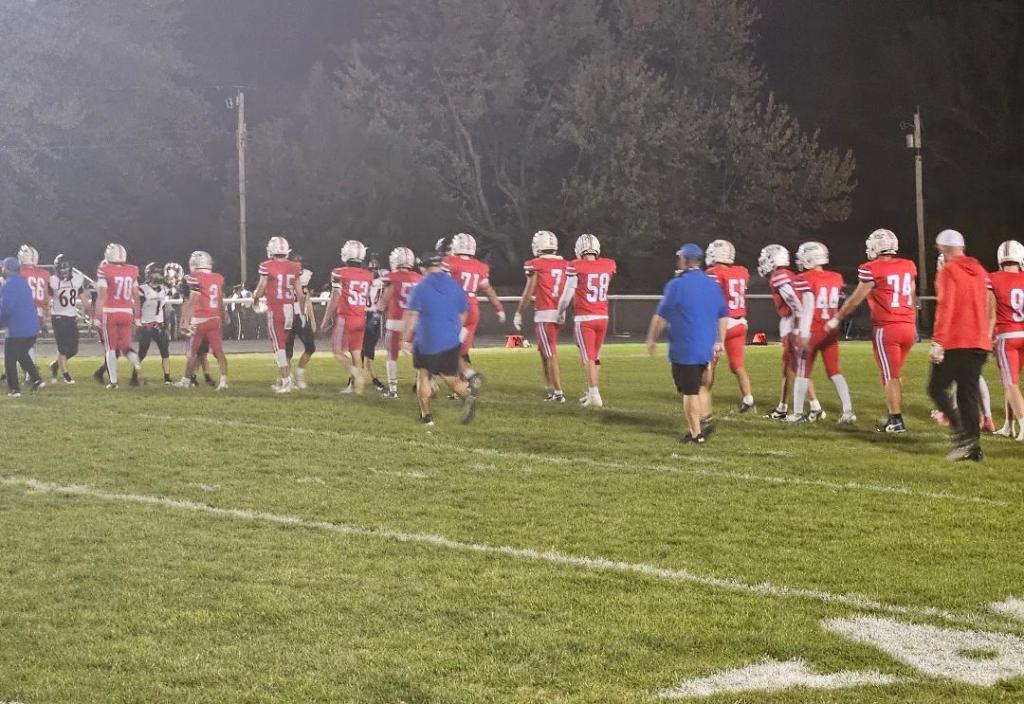 High school football players in red and white uniforms walk across the field after a game, shaking hands with opponents.
