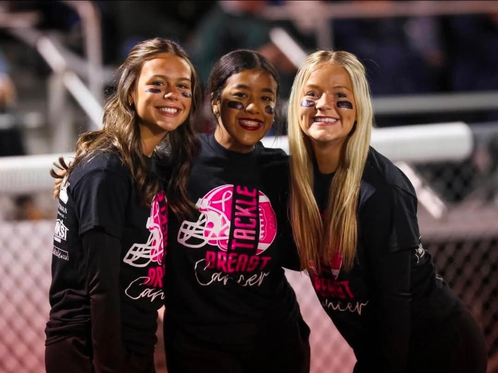 Three students at a football game, wearing matching black shirts with pink lettering supporting breast cancer awareness, smiling and posing for the camera.