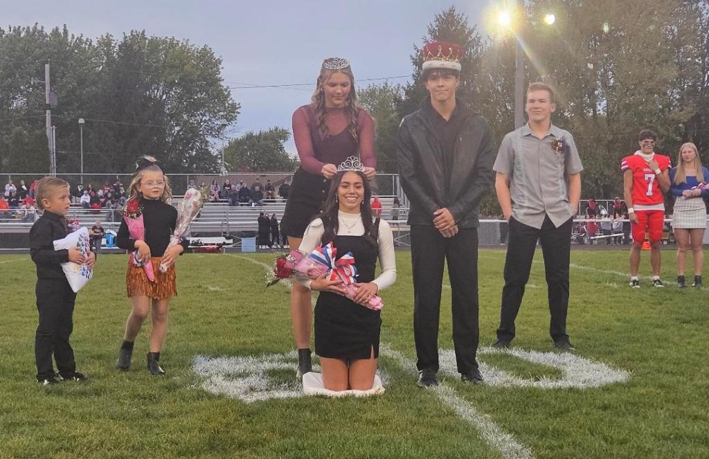 Ceremony crowning the homecoming king and queen, with crowned students standing on a football field surrounded by peers and young helpers.