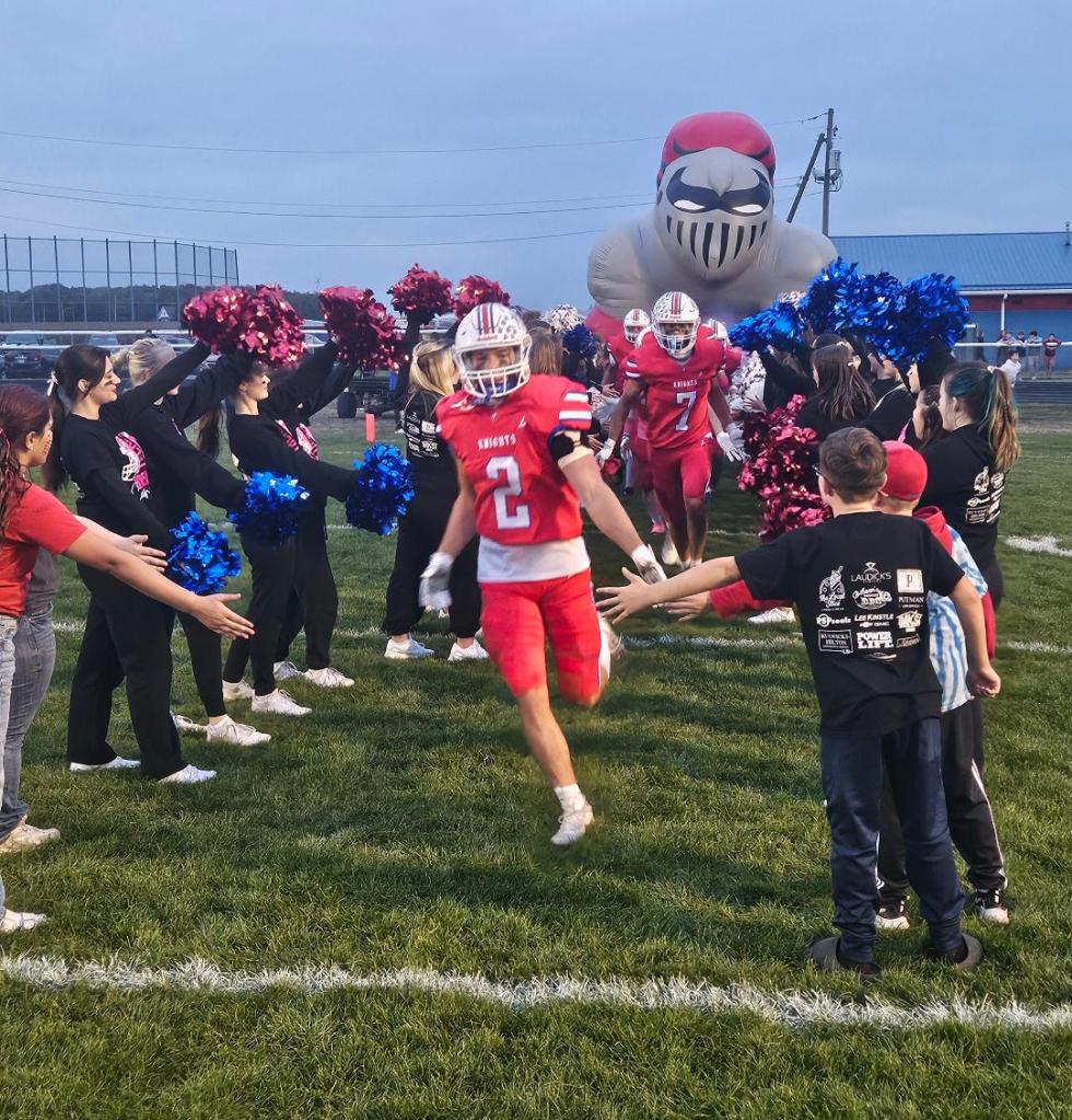 A football player wearing a red jersey with the number 2 runs through a cheerleader tunnel, with cheerleaders in black and colorful pom-poms on either side, as a large inflatable mascot looms in the background.
