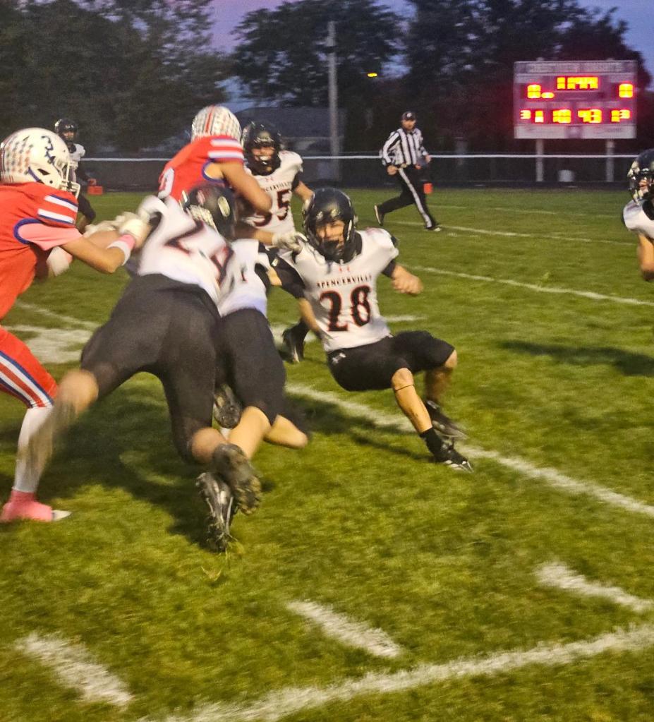 A football player in a black jersey is attempting to tackle an opponent in a red jersey while other players are engaged in the play on a grass field under dim lighting.